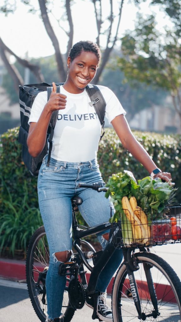 Smiling African American woman on bicycle delivering groceries outdoors, showing thumbs up.
